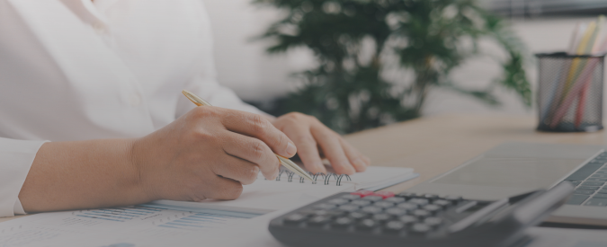 Image shows a hand holding a pen, pieces of paper, a calculator and a laptop