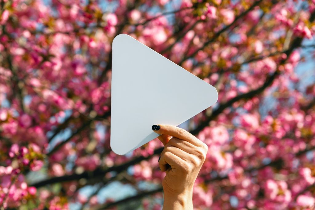 Hand holding a white play button icon in front of blooming cherry blossom tree on a bright spring day.