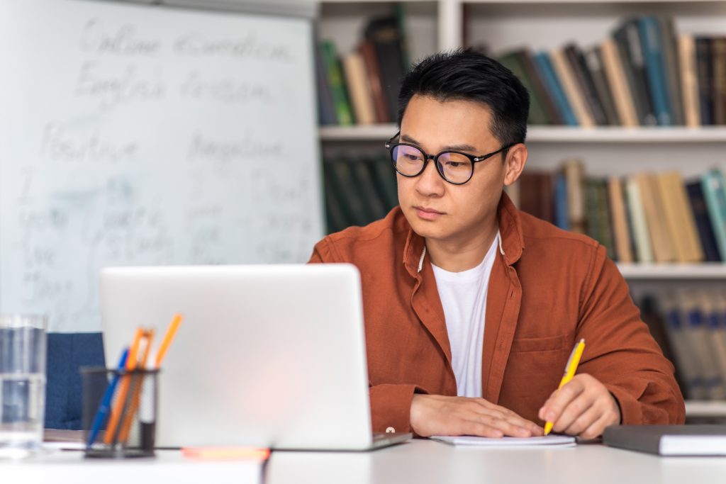 E-Teaching. Korean Middle Aged Man Teacher Using Laptop And Taking Notes Preparing For Virtual Lecture Sitting At Desk In Modern Classroom. Internet Technology And Education. Selective Focus