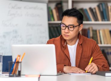 E-Teaching. Korean Middle Aged Man Teacher Using Laptop And Taking Notes Preparing For Virtual Lecture Sitting At Desk In Modern Classroom. Internet Technology And Education. Selective Focus