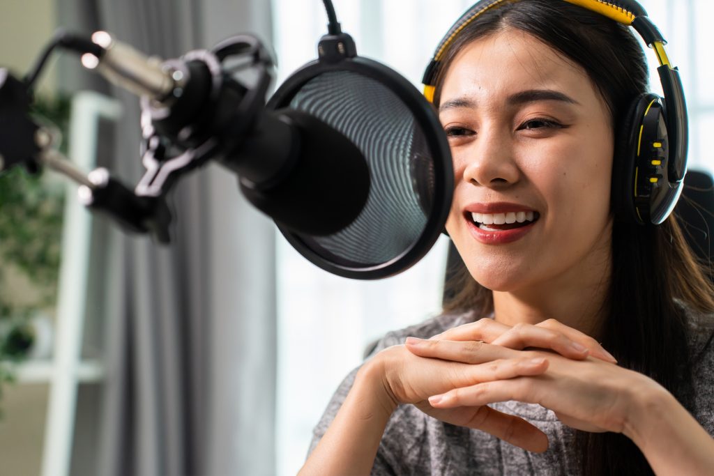 Asian attractive audio DJ woman speaks into microphone to broadcasting. Young beautiful female blogger influencer wearing headphones and recording morning news podcast show for radio at home studio.